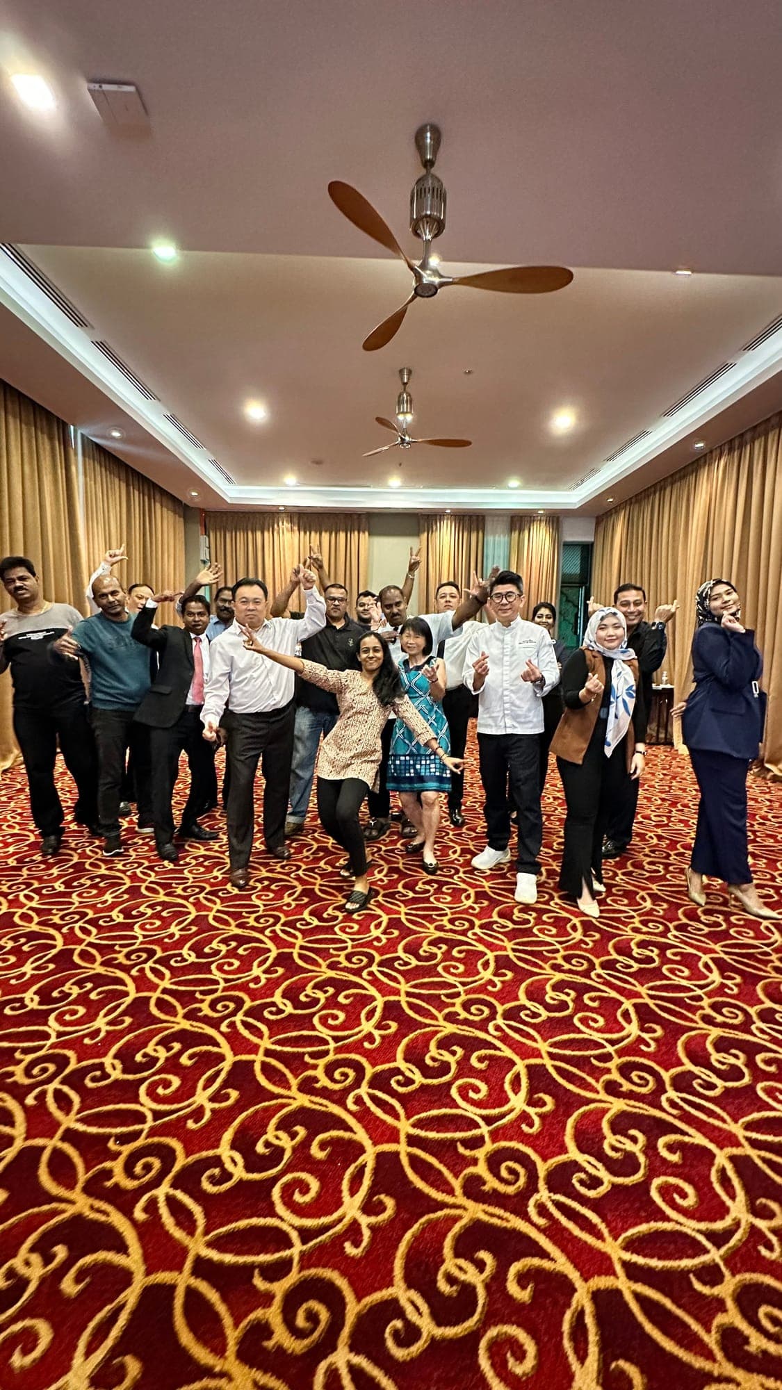 A large group of colleagues dancing together in a hotel ballroom with a red patterned carpet