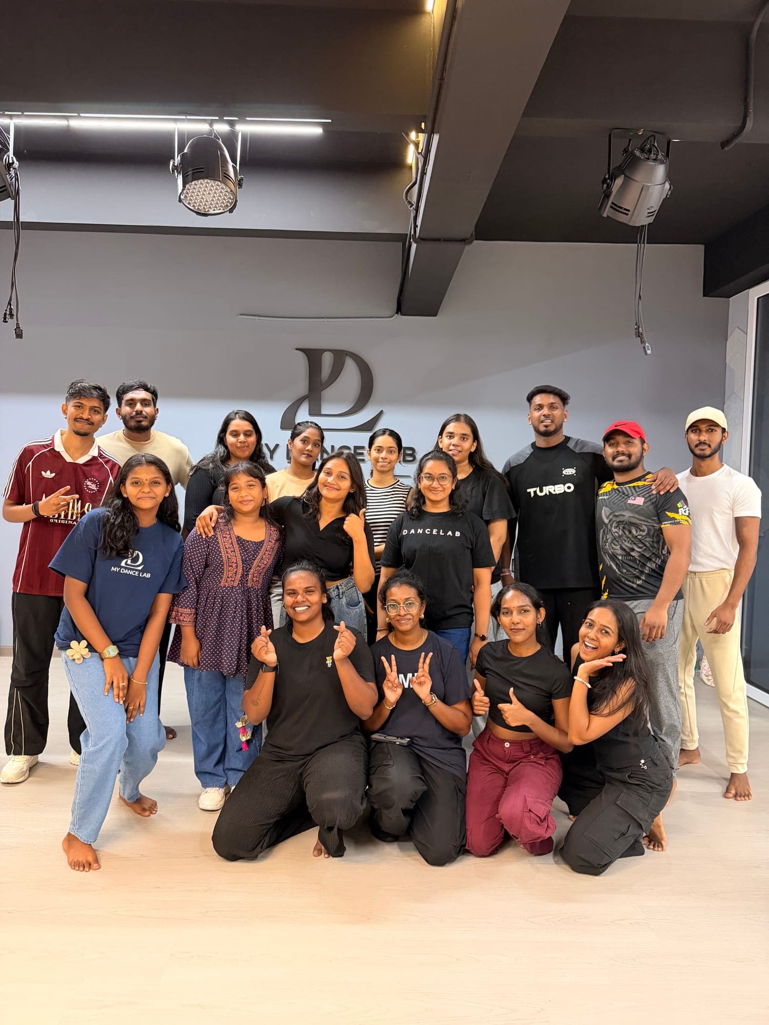 A smiling group of dancers, some in My Dance Lab T-shirts, at a dance workshop in front of the studio logo