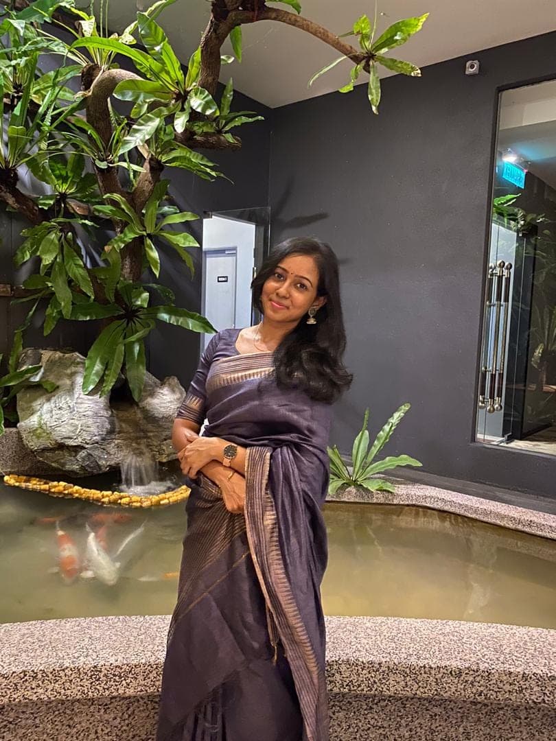 Hema, founder of My Dance Lab, smiling in a purple silk saree beside an indoor koi pond