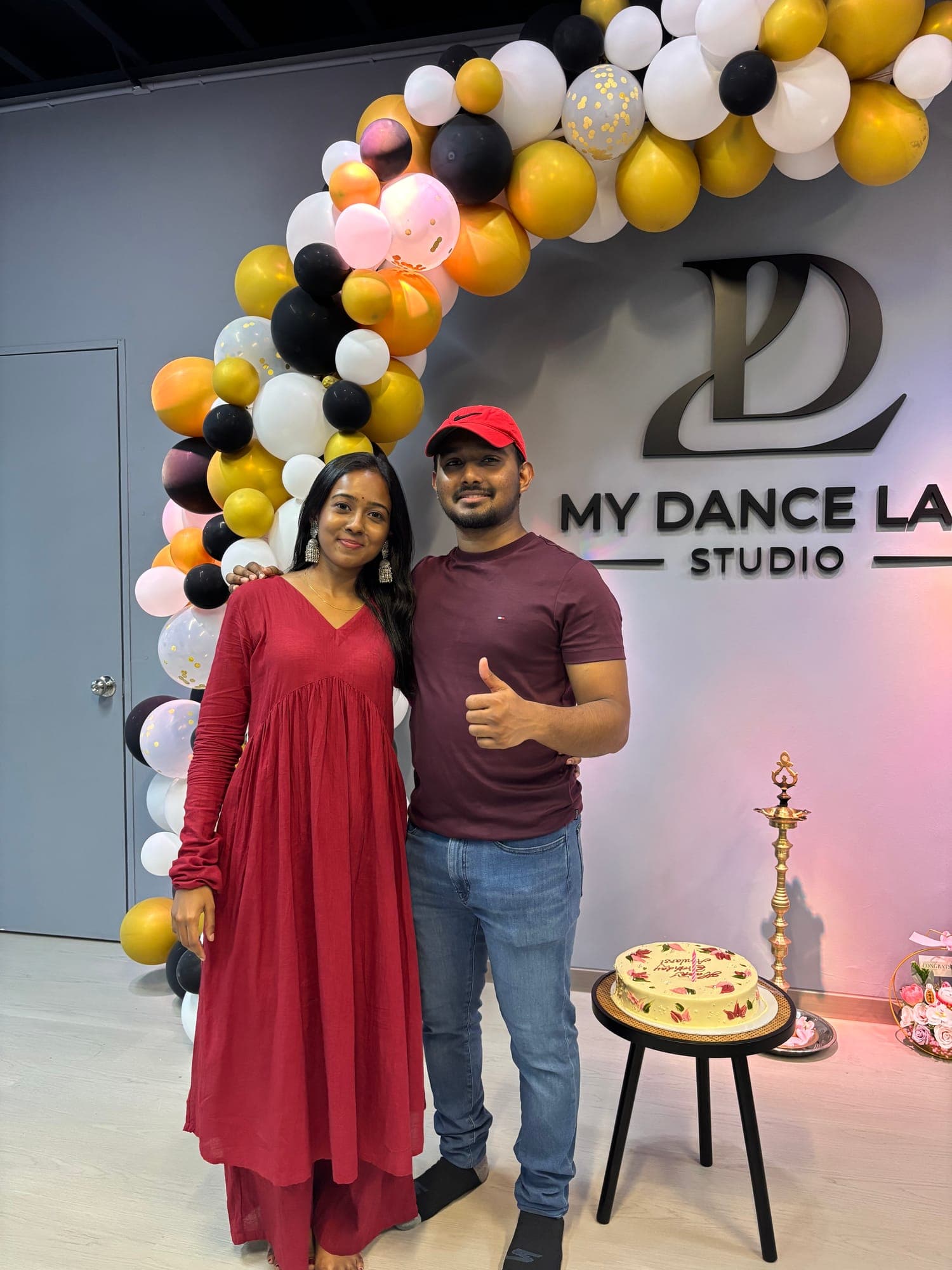 A woman in a red dress and a man in a red cap smiling beside the My Dance Lab Studio logo and a balloon arch on opening day