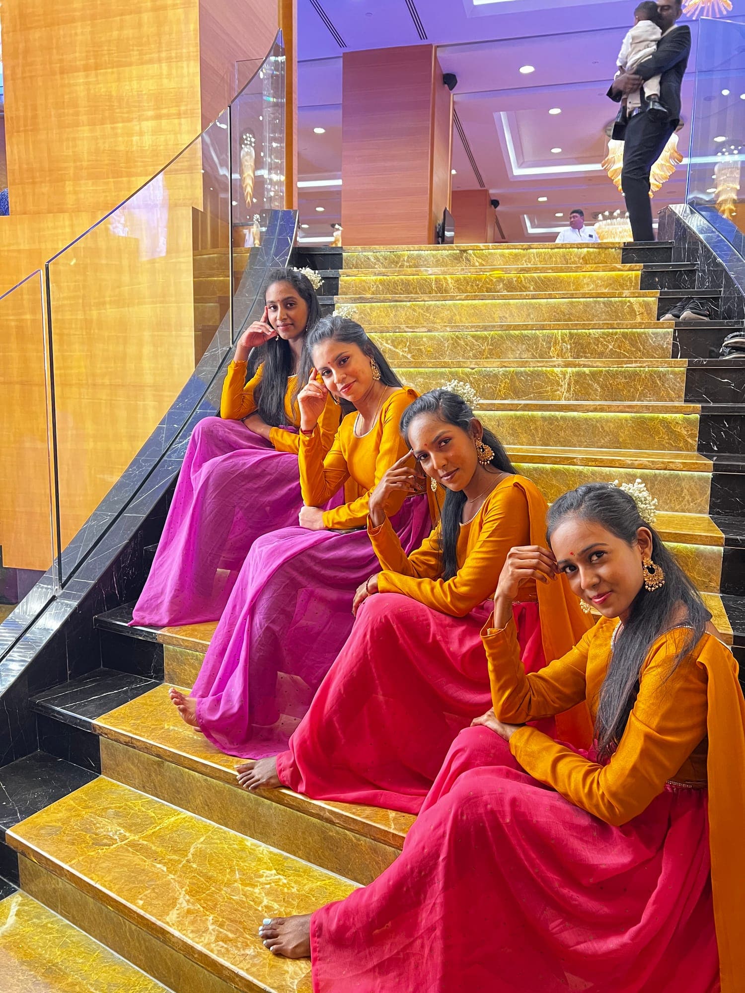 Four dancers in orange tops and pink skirts sitting on a golden staircase at an event venue