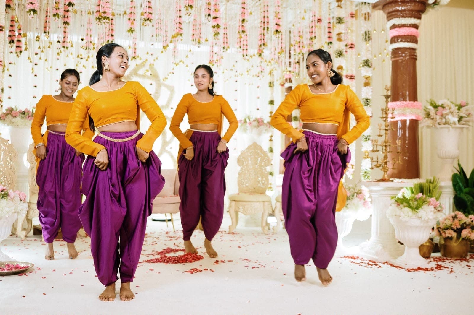Four dancers in mustard tops and purple dhoti pants performing on a stage decorated with flowers
