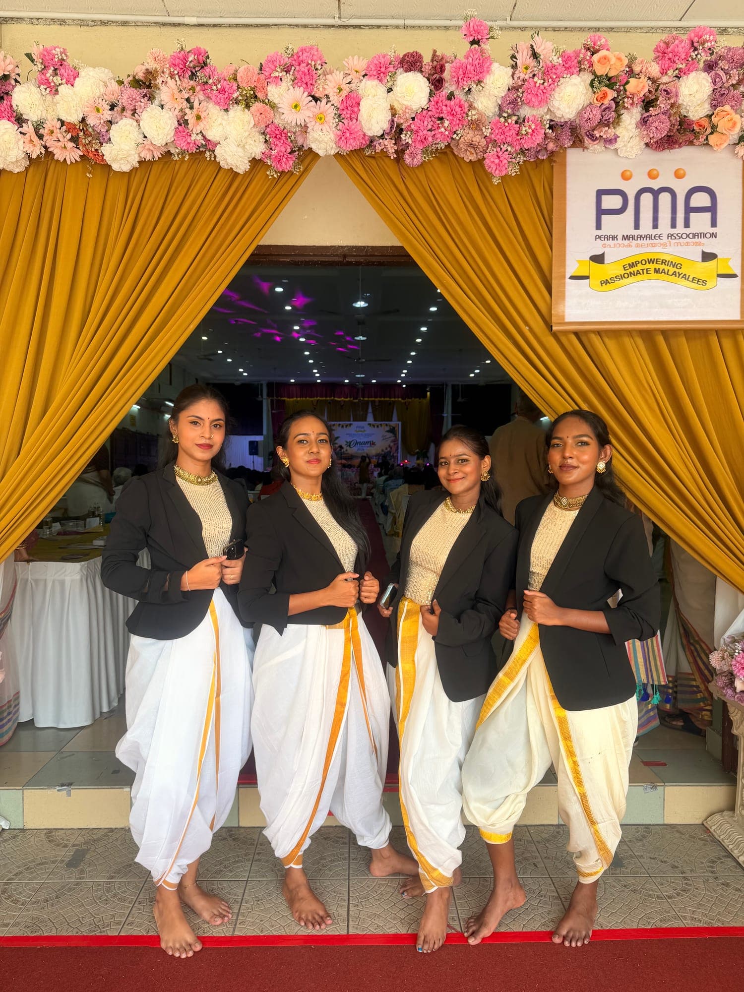 Four dancers in black jackets and white dhoti pants at an event entrance decorated with flowers