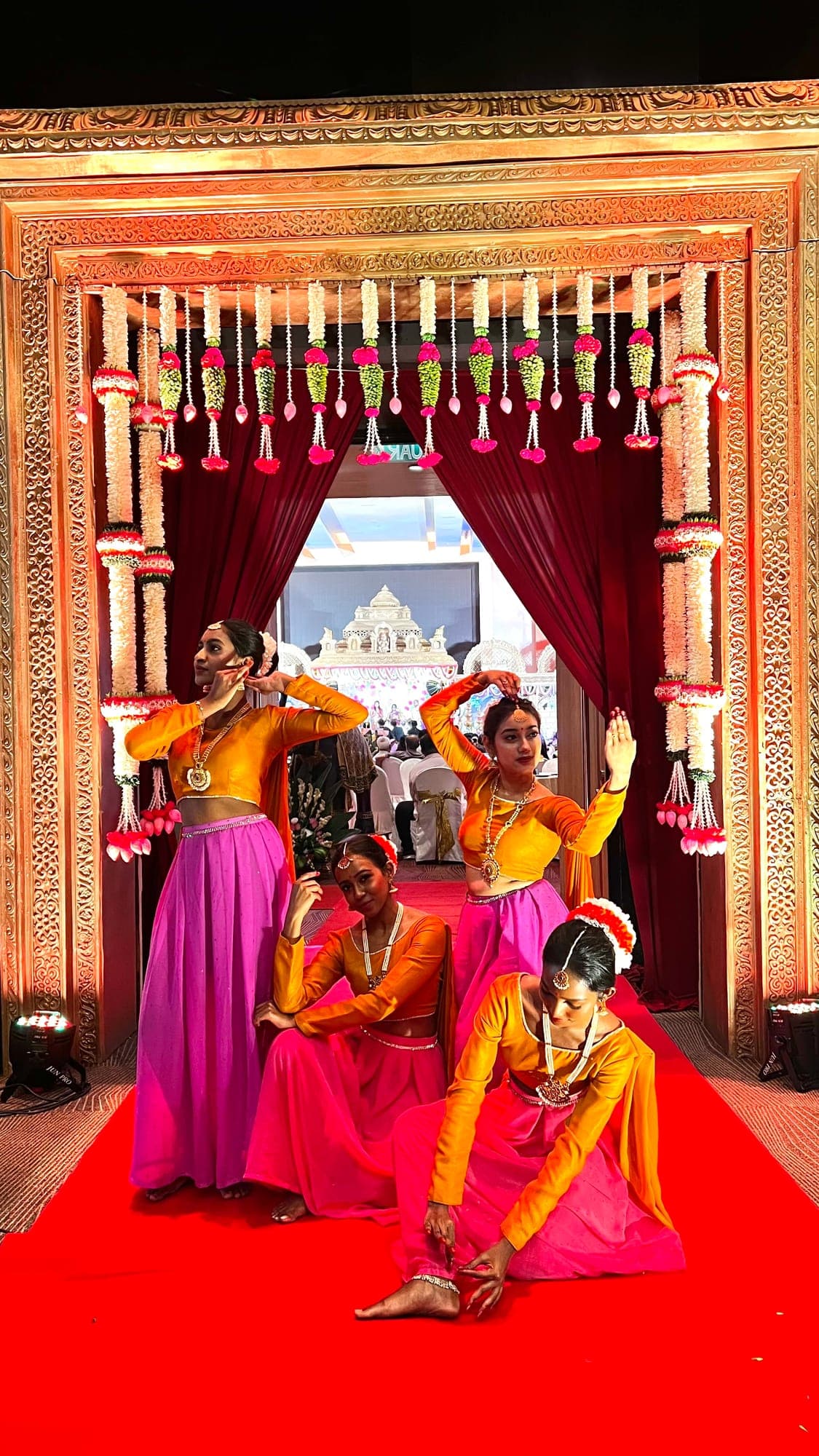 Dancers in orange tops and pink skirts posing on a red carpet beneath an ornate floral archway